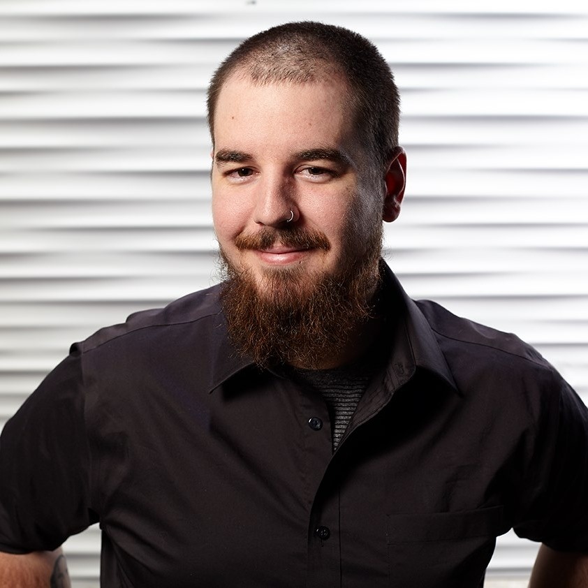 A professional headshot of Sebastian Inman, the founder of BetterA11y. He is a man with short hair, a beard, and a nose ring, smiling confidently with his hands on his hips. He wears a black button-down shirt against a textured white background.